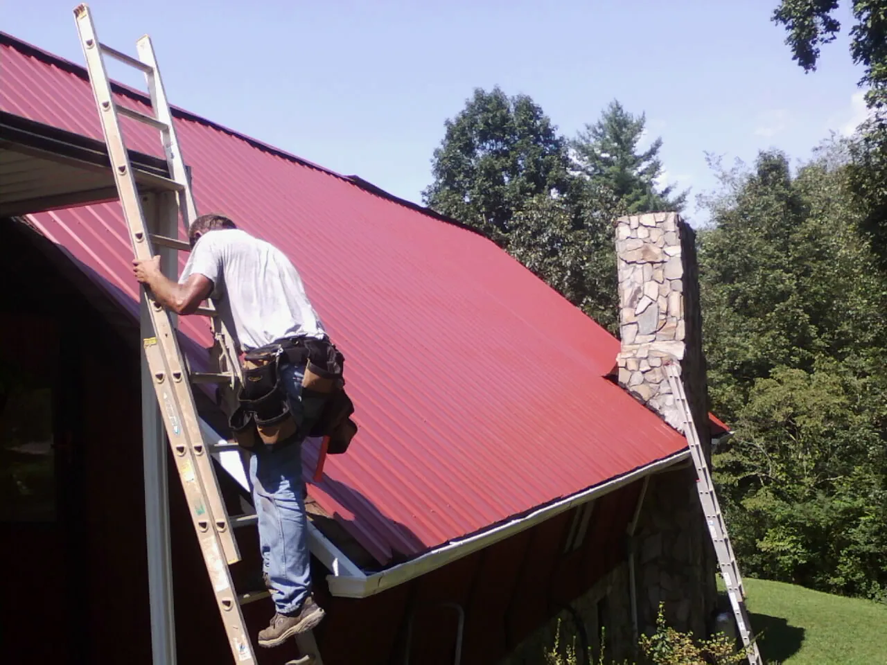 Roofer on a ladder beside a red metal roof and stone chimney — Soffit & Fascia Repair by Roofing Brothers in New Troy